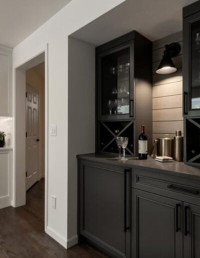 A modern kitchen with dark wood flooring, white cabinetry, and a built-in dark gray bar area stocked with wine bottles and glasses, illuminated by a wall-mounted light.