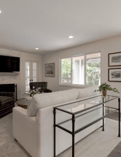 A modern living room features a large sofa, two plaid armchairs, a glass coffee table, a wall-mounted TV above a fireplace, and multiple windows allowing natural light to enter.