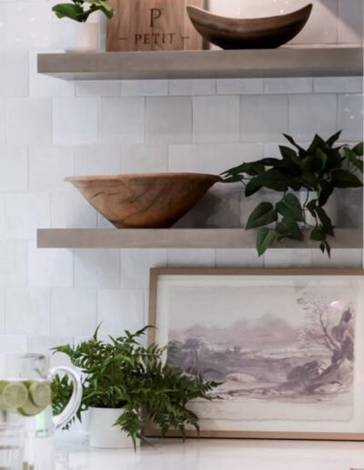 Two wooden shelves on a white-tiled wall display a potted plant, wooden bowls, and a cutting board. Below the shelves, a framed landscape artwork and fern plant rest on a white countertop.