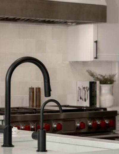 A modern kitchen with a black faucet on a white island countertop, a stove with red knobs in the background, and white cabinets above.