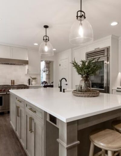 A modern kitchen with white cabinets, stainless steel appliances, a large island with bar stools, pendant lights, and hardwood floors.