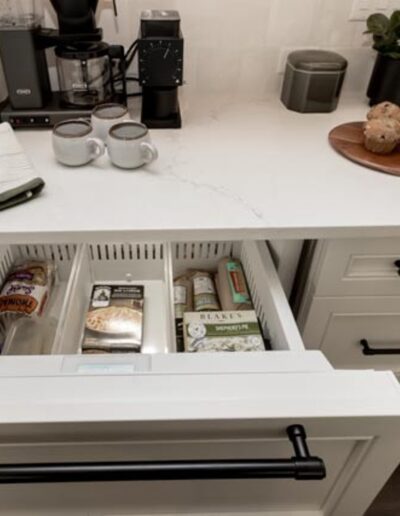 A kitchen counter with a coffee maker, two mugs, a towel, and a tray of muffins. Below, a drawer is open revealing various food items including bread and snacks.