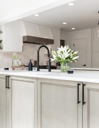 A modern, well-lit kitchen featuring a large island with white countertops, a black faucet, flowers, and minimalist decor. The background includes white cabinetry, stainless steel appliances, and shelves.