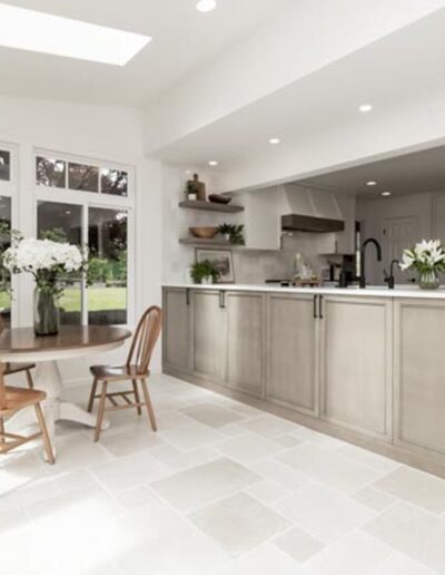 A modern kitchen with light-colored cabinets, a central island, and a dining area with a round wooden table and two chairs. The space is bright with large windows and a skylight.