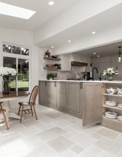 A bright kitchen and dining area with wooden cabinets, a small round table, chairs, pendant lighting, and open shelves displaying white crockery. Sunlight streams through large windows.