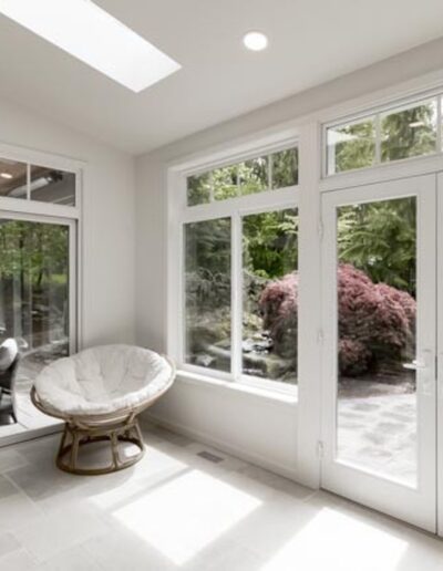 A bright sunroom with large windows and a glass door opens to a lush outdoor area. Inside, there's a round cushioned chair facing the windows.