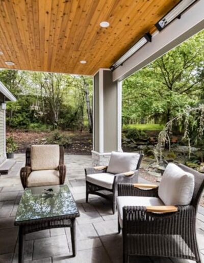 Outdoor patio with a wooden ceiling, cushioned seating, glass-top coffee table, and wicker chairs, adjacent to a natural backyard with greenery and a small pond.