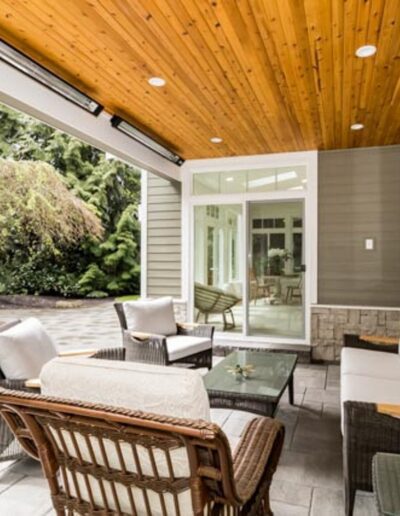 A covered patio with a wooden ceiling, white cushioned wicker seating, a glass-top coffee table, and a view of a landscaped garden with a small pond.